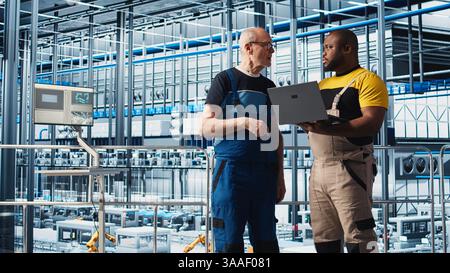 Senior engineer in smart photovoltaics factory talks with intern, showing system performance metrics. Experienced solar panels plant expert guiding new trainee in tracking energy production, camera A Stock Photo