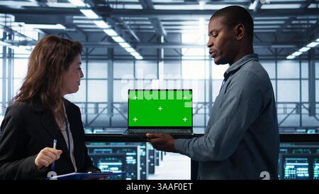 Technicians in data center using green screen mockup laptop to develop disaster recovery plans. IT specialists in server farm testing failover scenarios on chroma key device, protecting data, camera B Stock Photo