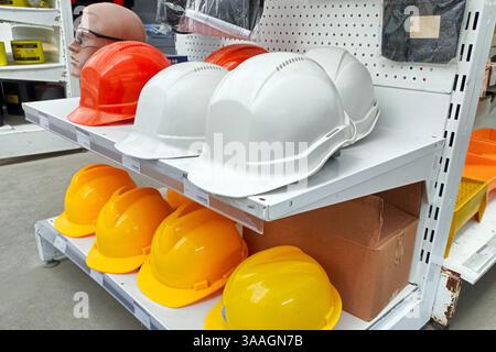 A variety of construction safety helmets in white, orange, and yellow displayed on store shelves, with a mannequin head wearing glasses. Stock Photo
