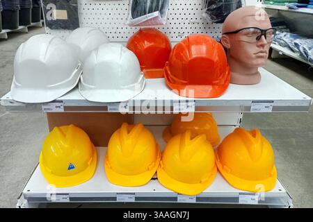 A variety of construction safety helmets in white, orange, and yellow displayed on store shelves, with a mannequin head wearing glasses. Stock Photo