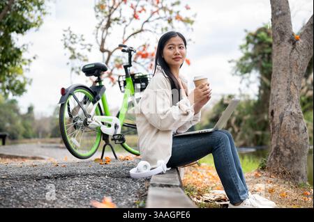 An asian woman is sitting on the sidewalk and holding a drink with both hand, taking a break from cycling in the park or garden. relaxing in the natur Stock Photo
