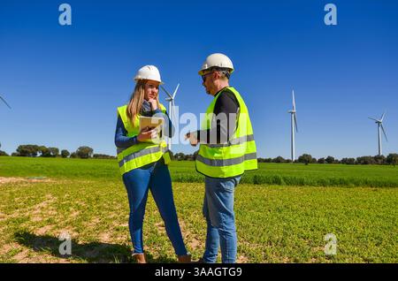 Two engineers wearing safety vests and helmets are discussing using a tablet next to wind turbines in a green field, promoting renewable energy and su Stock Photo