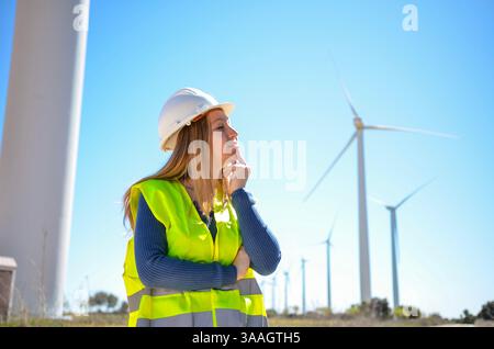 Confident female engineer wearing safety vest and helmet thinking about next renewable energy project at a wind turbine power plant Stock Photo