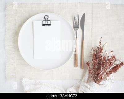 Romantic table plate setting in light color, empty plate with white paper for the menu, fork and knife on linen tablecloth and dry flowers Stock Photo