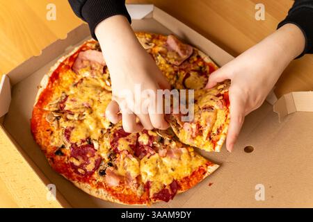 Two hands are pulling apart a freshly baked pizza topped with cheese, meats, and vegetables. The pizza is placed inside an open takeout box on a woode Stock Photo