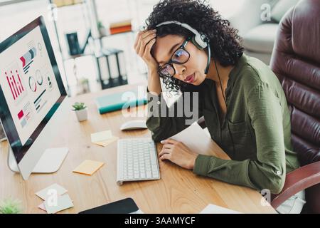 Young female professional working in office, wearing headset and analyzing data on computer with a thoughtful expression and casual attire Stock Photo