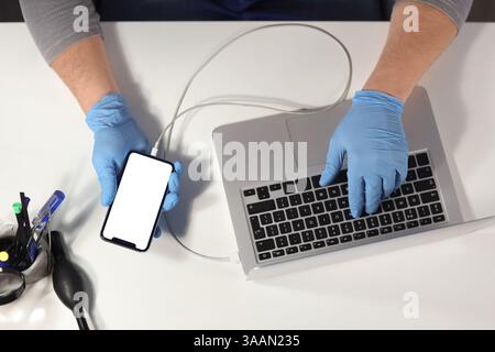 Male technician checking mobile phone with laptop on table in service center, top view Stock Photo