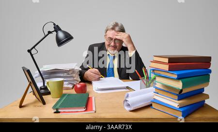 Elderly man, professor sitting at desk with many books and supplies, checking students papers with little smile Stock Photo