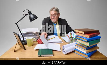 Senior man checking documents at cluttered desk with skeptical expression against light grey background. Stock Photo