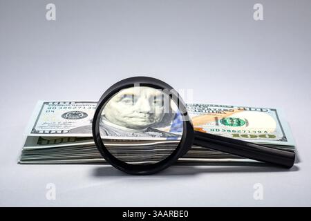 A magnifying glass focuses on a stack of hundred dollar bills, highlighting the intricate details of the currency and emphasizing its important role in economic transactions. Stock Photo