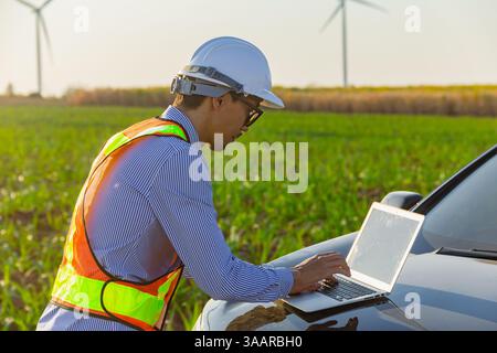 Professional Engineer young man working on laptop at front of car hood countryside outdoor with wind turbine farm background Stock Photo