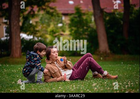 Smiling child watches as parent blows bubbles while relaxing on green grass in a park. Stock Photo