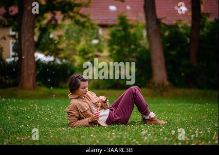 Person lies on green grass blowing soap bubbles, enjoying a peaceful moment outdoors. Stock Photo