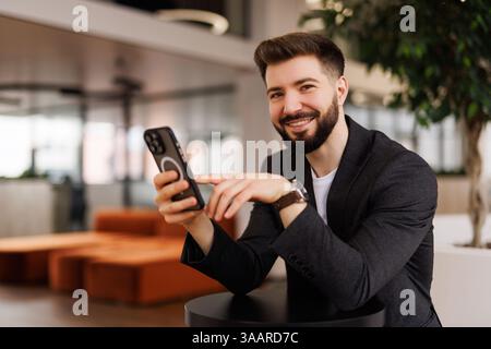 A smiling young male office worker sits in the office at a desk and uses a mobile phone, writes a message, corresponds. Stock Photo
