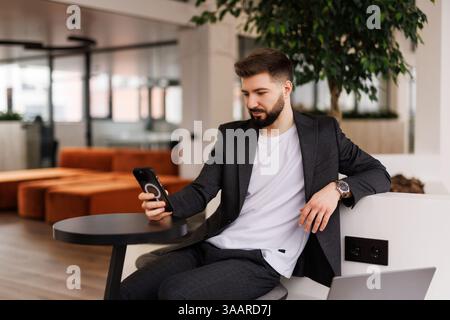 A smiling young male office worker sits in the office at a desk and uses a mobile phone, writes a message, corresponds. Stock Photo
