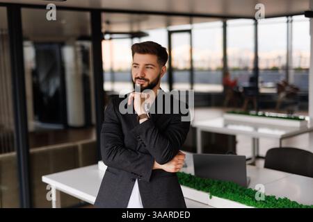 Serious confident business man, thoughtful doubtful company ceo executive wearing blue suit standing in office holding hand in chin looking at camera Stock Photo
