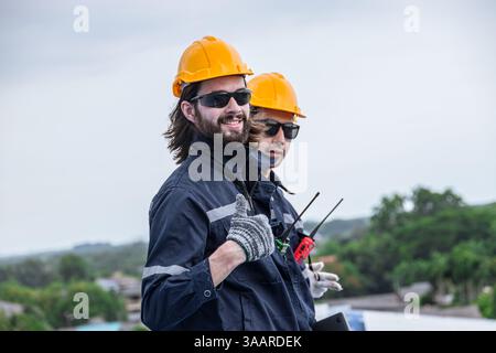 Happy engineer worker team standing thumbs up wearing groove safety suit hardhat work at height danger place roof top outdoors Stock Photo