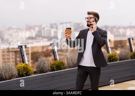 Take a pause. Waist up portrait of smiling businessman drinking coffee on office balcony after talking by phone. Copy space on right Stock Photo