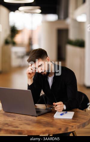 Pensive young manager man working at laptop, sitting at workplace table, looking at display, typing, touching chin, thinking on online content, using Stock Photo