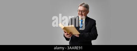 Senior academic man in suit smiling while reading hardcover book on grey background. Stock Photo