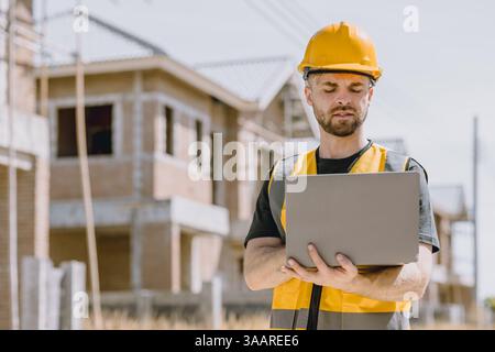 Construction worker Engineer male in safety vest and helmet looking laptop computer project management software at construction site Stock Photo