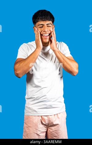 Young hispanic man holding face with hands and laughing on blue background Stock Photo