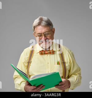 Elderly man smiling while reading book, enjoying academic material on neutral background. Calm moment, love for reading, daily work joy. Stock Photo