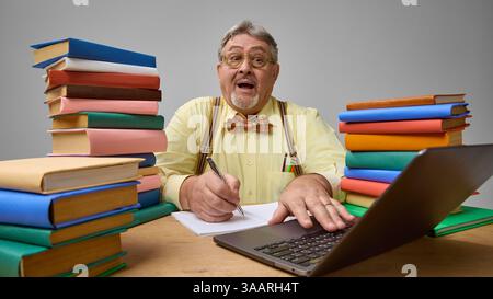 Elderly man, scholar expressing surprise while writing in notebook and working on laptop. Stock Photo