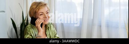 Mature woman enjoys chatting on the phone in a bright, airy room filled with natural light. Stock Photo