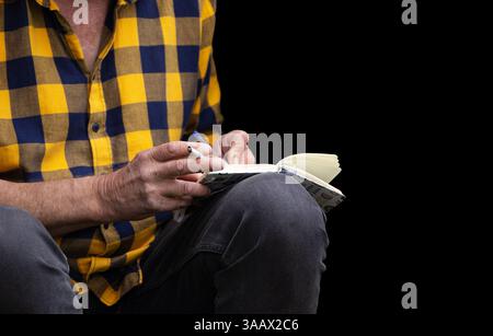 Closeup of a man in a yellow  checkered shirt holding a pen and a cigarette, writing in notebook Smoking habits, creativity, note taking, journaling, Stock Photo