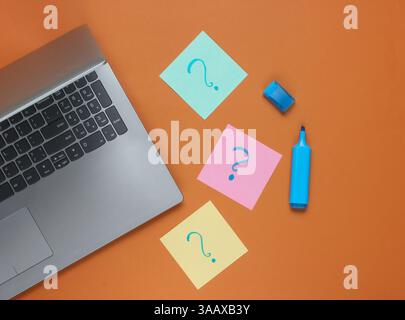 Laptop, memo pieces of paper with drawn question marks and felt-tip pen on brown background. Top view Stock Photo