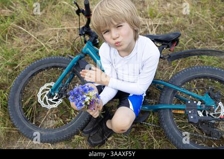 boy is relaxing sitting on bicycle, holding small bouquet of wild flowers in hands as gift for mom. Healthy outdoor activity. Summer day on holiday in Stock Photo