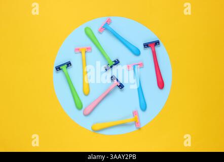 Many colored plastic razors on yellow background with blue pastel circle. Minimalistic beauty still life Stock Photo