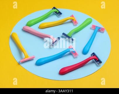 Many colored plastic razors on yellow background with blue pastel circle. Minimalistic beauty still life Stock Photo