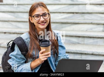 Happy modern woman student in denim jacket and backpack sitting on the stairs with laptop outdoor. Drinks coffee. Distance learning. Modern youth conc Stock Photo