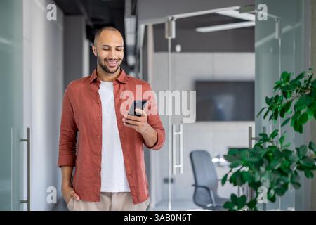 Successful businessman walking through the office with a phone in his hands, the man is typing a text message and smiling, browsing social networks and internet pages at his workplace. Stock Photo