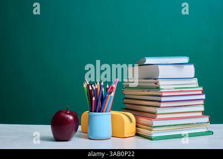 Stack of colorful books, a mug filled with colored pencils, an apple, and a yellow pencil case are arranged on a desk against a green chalkboard backg Stock Photo