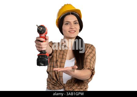 Confident female construction worker in a yellow helmet holding a cordless drill forward, smiling and presenting it with an open hand, isolated on whi Stock Photo