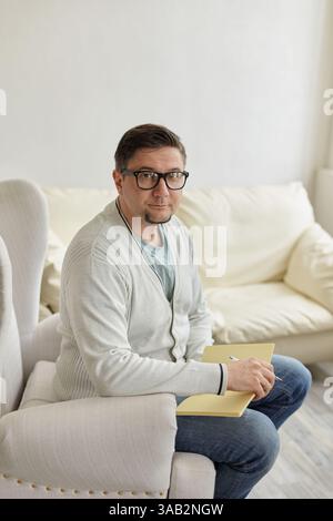 Portrait of handsome male psychologist and taking notes during therapy session at clinic. Friendly millennial psychotherapist posing and smiling at of Stock Photo