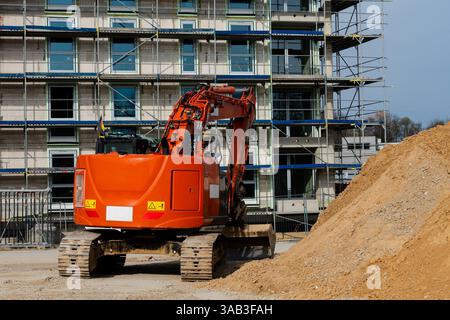 An orange excavator is operating on a construction site, moving dirt and debris near a partially built structure surrounded by scaffolding. Workers ar Stock Photo