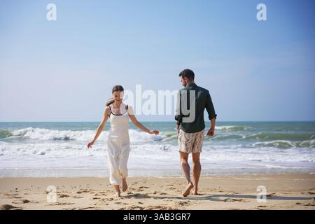 A carefree young couple enjoys a relaxing walk on a sandy beach, barefoot and smiling under the clear sky. Gentle ocean waves roll in behind them, creating a tranquil and joyful moment full of summer Stock Photo