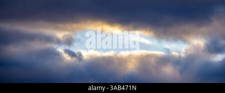 A dramatic sky with dark clouds partially revealing a patch of blue sky and sunlight. Stock Photo