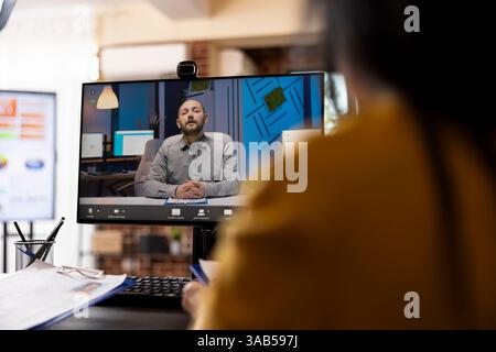 Selective focus on a computer screen displaying a video call with a caucasian businessman. Rearview closeup of an individual attending virtual meeting with a white male company consultant. Stock Photo