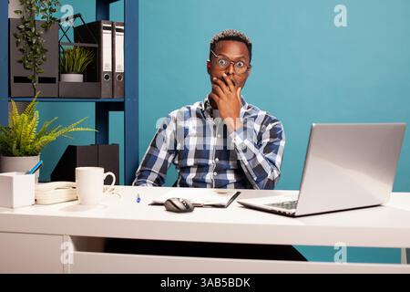 African american manager with hand on jaw, having a shocked expression, eyes wide open looking at camera. Portrait of a stunned black male employer cupping his mouth after reading a surprising email. Stock Photo