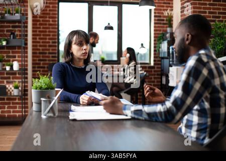 Caucasian woman shares her professional background with an African American HR employee. Male recruiter listens attentively to female job candidate in ongoing interview meeting at modern workplace. Stock Photo