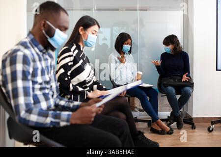 Young diverse women in face masks, sitting in waiting area and talking about possible job offers at startup company. Multiethnic people chatting and discussing interview process in office lobby. Stock Photo