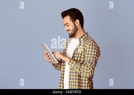 Handsome Indian guy using tablet computer for online work or communication on violet background Stock Photo