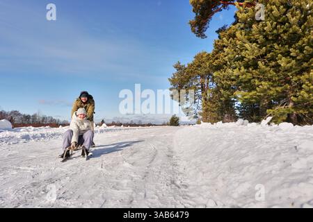 Riding wooden sled, woman sits as man pushes her across snowy path, mature European couple enjoy winter activity and having fun outdoors in nature. Stock Photo