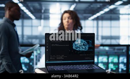 Laptop in front of data center engineers in blurry background deploying artificial intelligence driven management tools. AI driven notebook used for overseeing server clusters, camera B close up Stock Photo