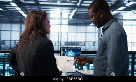 Data center team reviewing paperwork in server room, analyzing system reports using AI on tablet. Artificial intelligence server room admins looking over financial documents, camera B Stock Photo